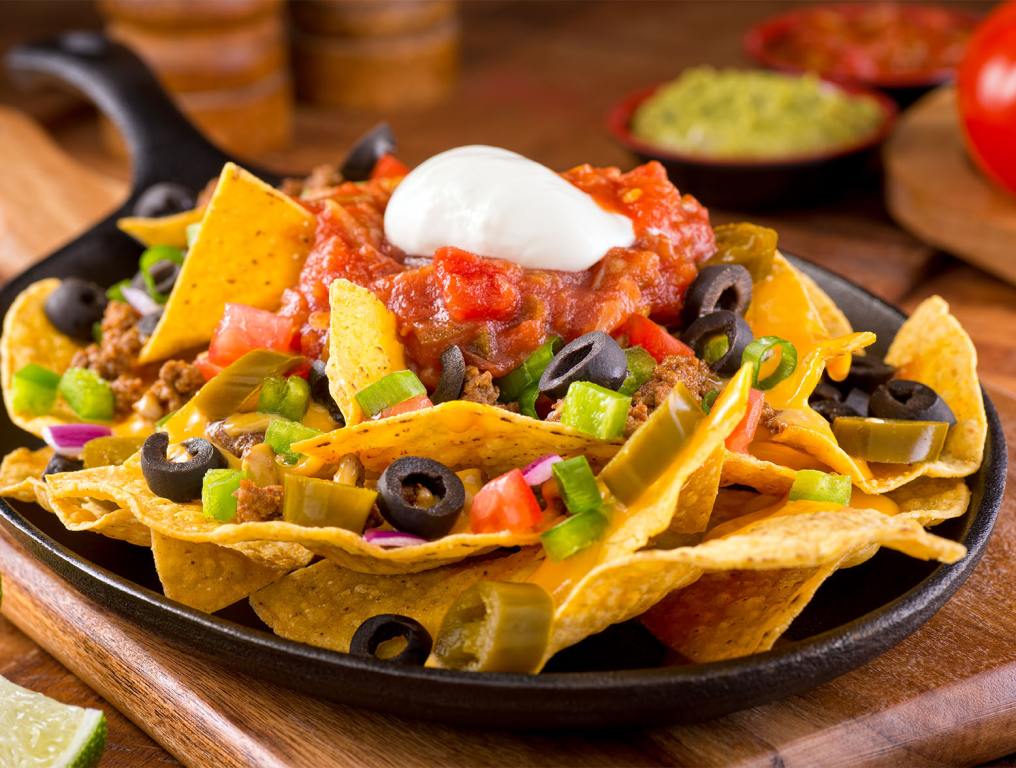 A plate of delicious tortilla nachos with melted cheese sauce, ground beef, jalapeno peppers, red onion, green onions, tomato, black olives, salsa, and sour cream with guacamole dip.